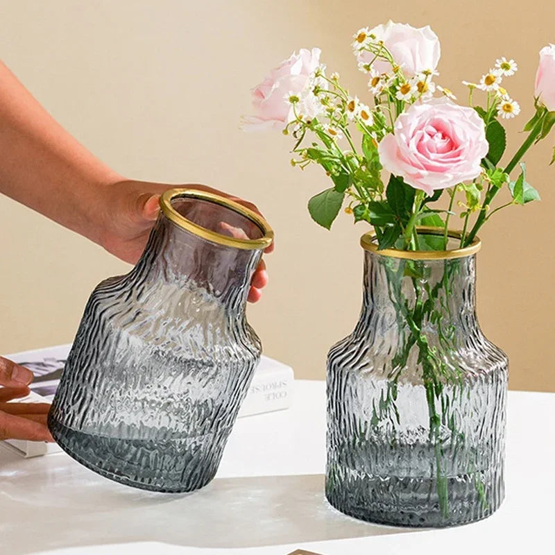 Clear glass vase with gold rim being held by a hand, filled with flowers on a light surface.