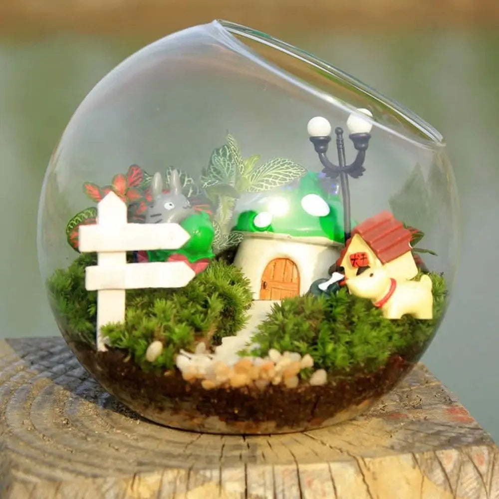 Glass terrarium with miniature landscape including a house, lamp post, and characters on a wooden surface.