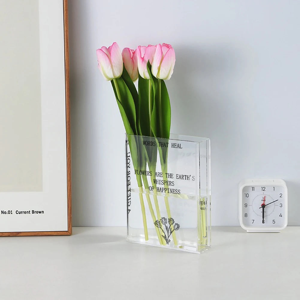 Clear vase with pink and white tulips on a white surface with a clock and framed picture in the background.