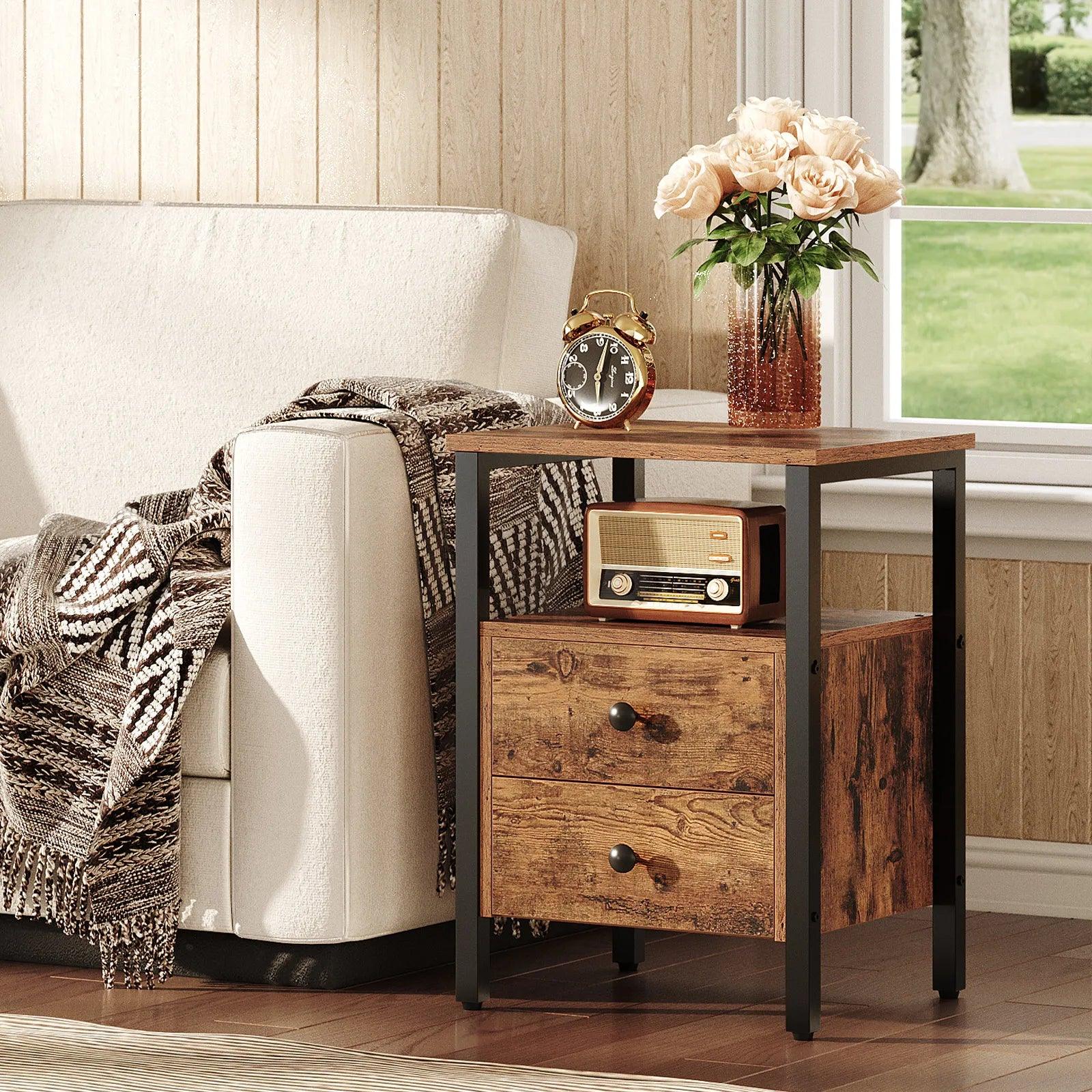 A rustic industrial nightstand with brown wooden top and black metal frame, featuring two drawers and an open shelf, placed next to a white sofa with a radio and a vase of flowers on top.