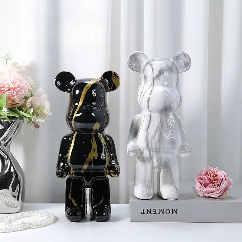 Two bear-shaped figurines, one black with gold accents and the other clear, on a white surface with flowers and a vase in the background.