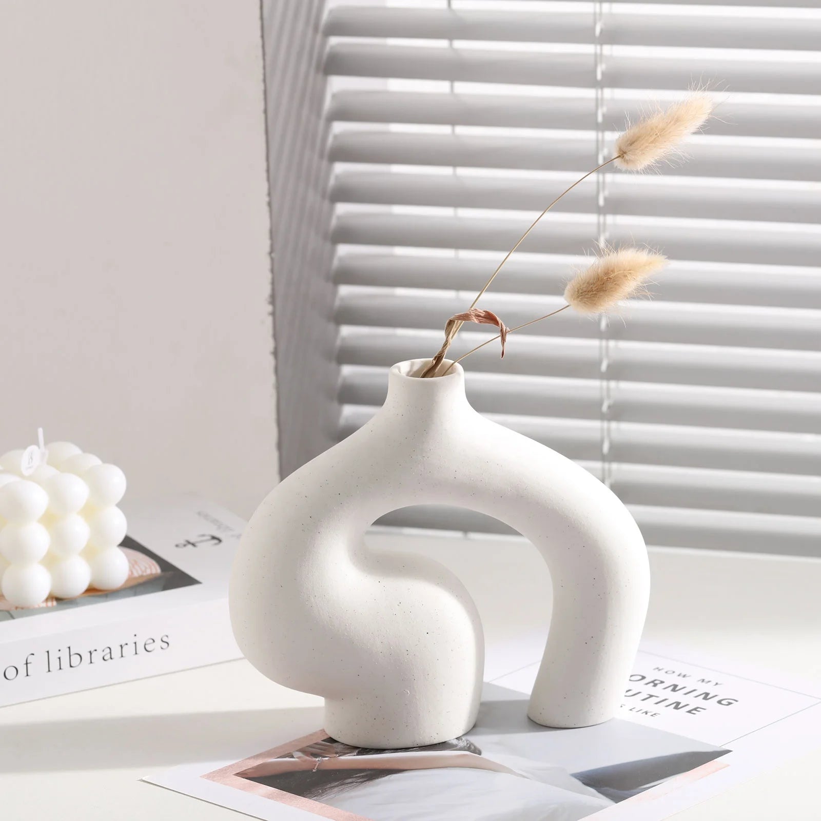 White ceramic vase with dried plant on a surface with books and decor items.