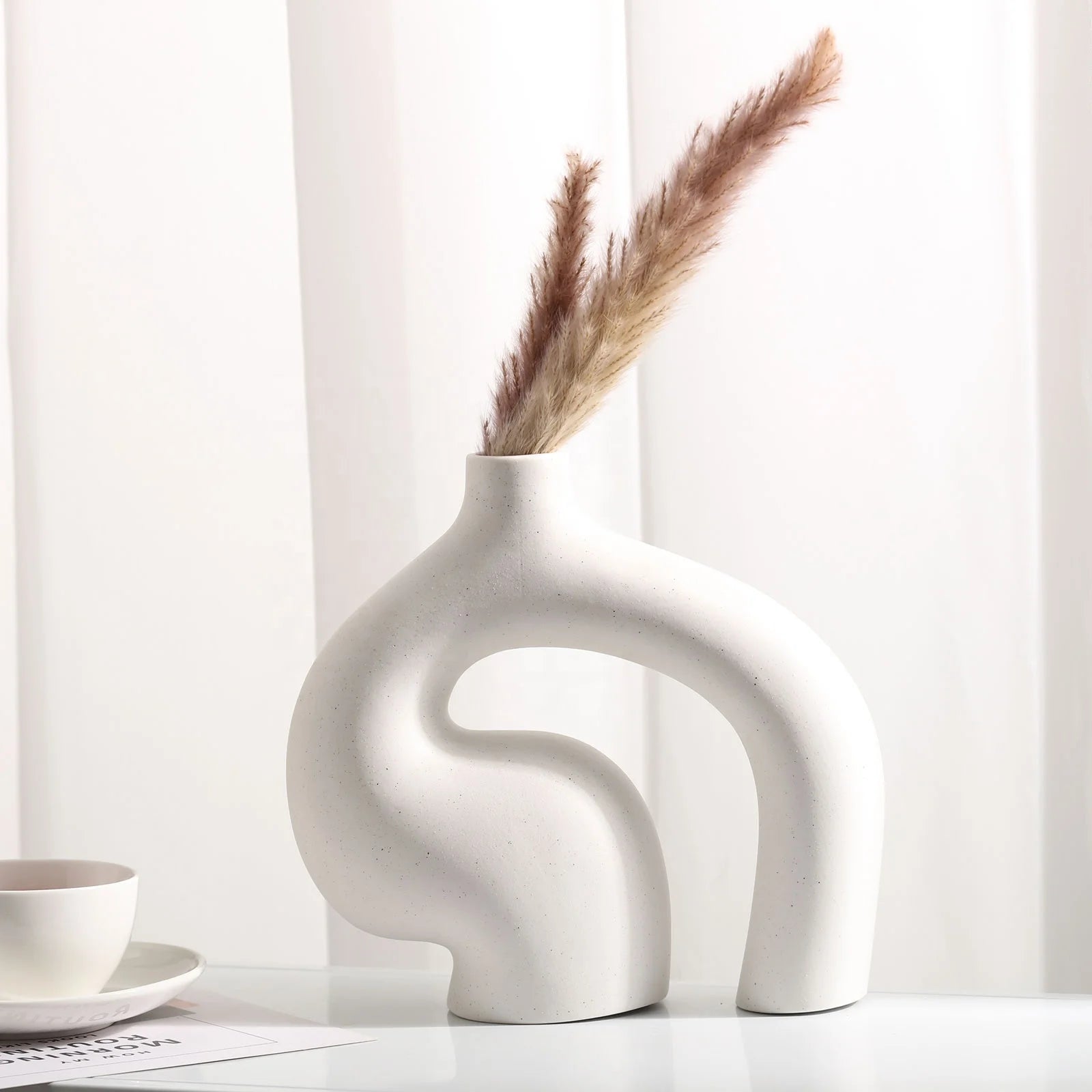 White abstract vase with dried pampas grass on a white surface with a white curtain background