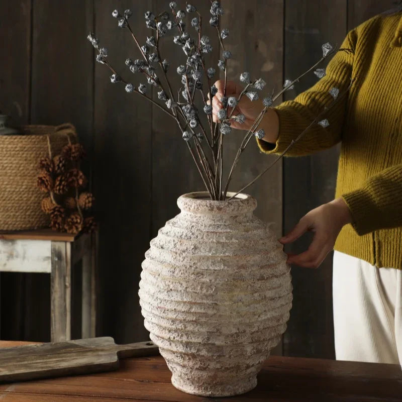 Person arranging dried branches into a textured vase on a wooden surface.