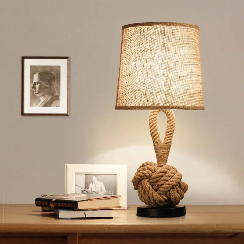 Table lamp with knotted base on a wooden surface, books, and framed pictures in the background.