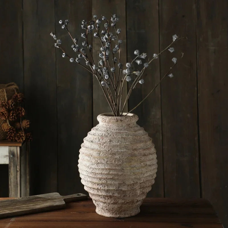 Textured vase with dried plants on a wooden surface against a dark wooden wall.