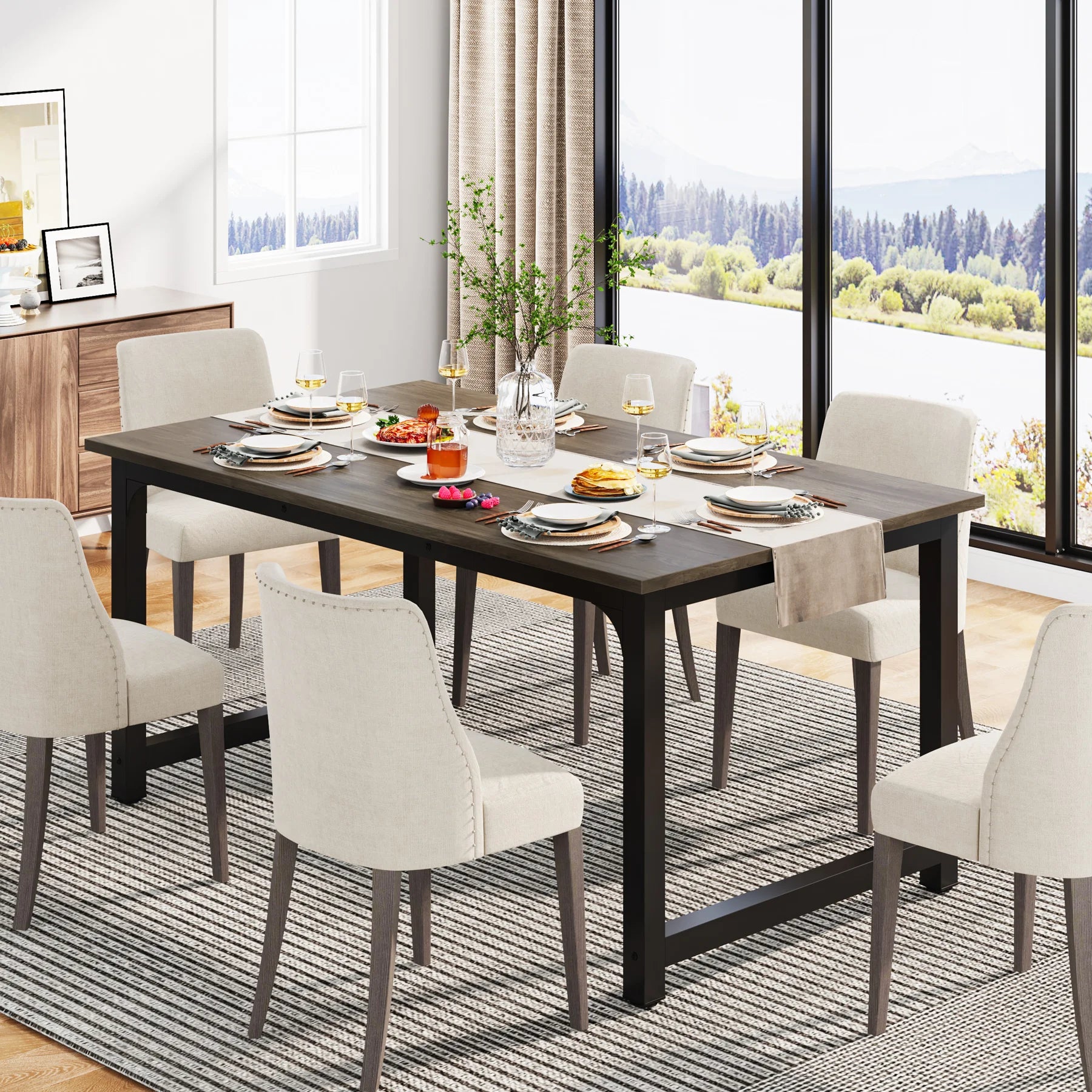 Dining room with a long dark wooden table set for a meal, surrounded by white chairs. Large windows show a scenic view.