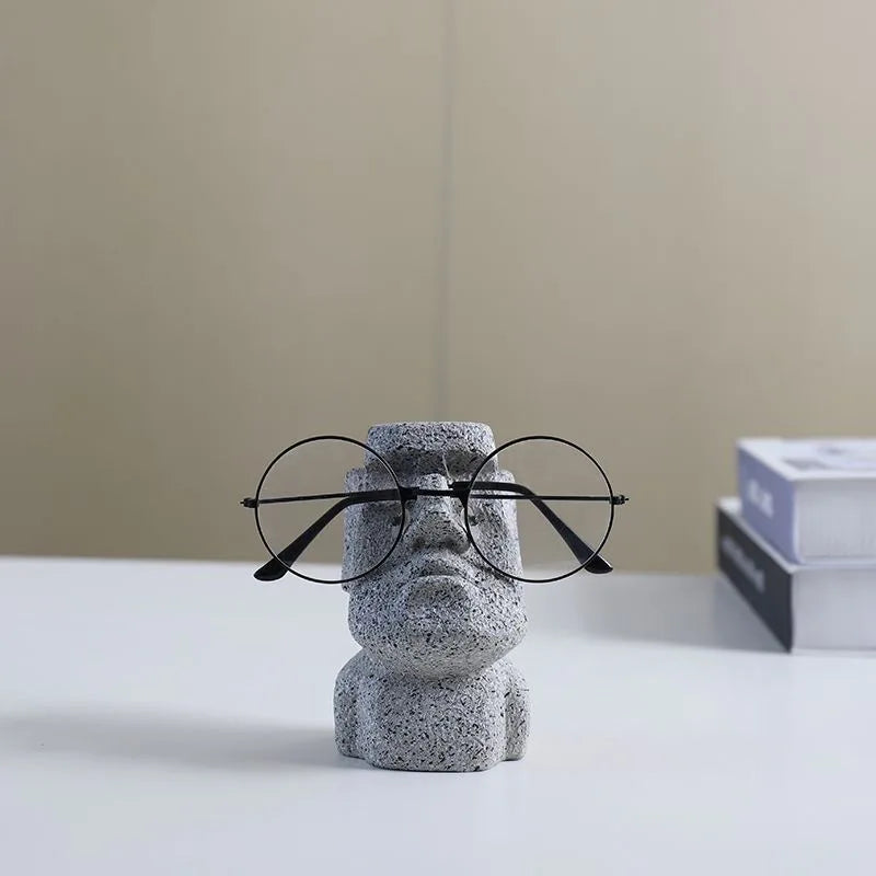 A resin figurine glasses holder with black frames, placed on a white surface, holding a pair of glasses in a stylized manner.