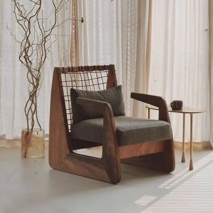 Modern armchair with wooden frame and grey cushion in a room with curtains and a small table.