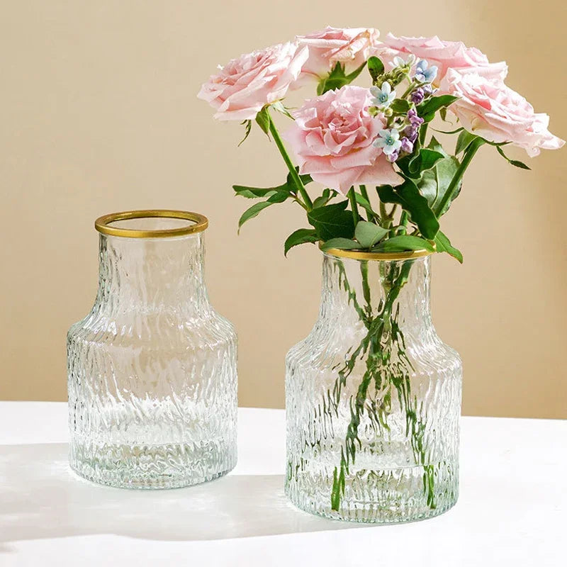 Two glass vases with gold rims, one containing pink flowers on a beige background.