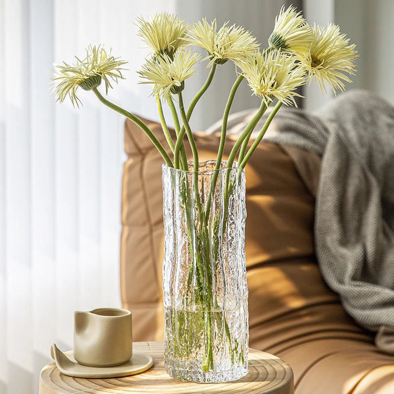 Clear glass vase with yellow flowers on a wooden table in a cozy living room setting.