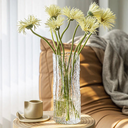 Clear glass vase with yellow flowers on a wooden table in a cozy living room setting.