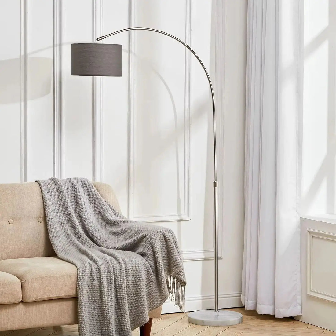 Floor lamp with a gray shade next to a beige sofa draped with a gray blanket in a room with white walls and curtains.
