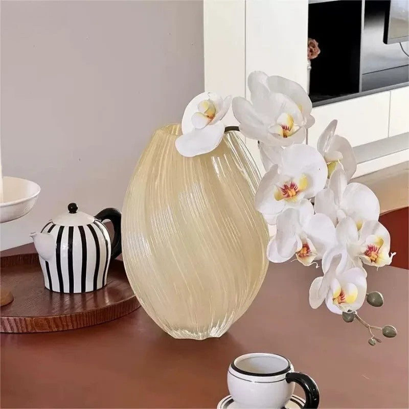 Beige vase with white orchids on a wooden surface with a teapot and cup in the background.