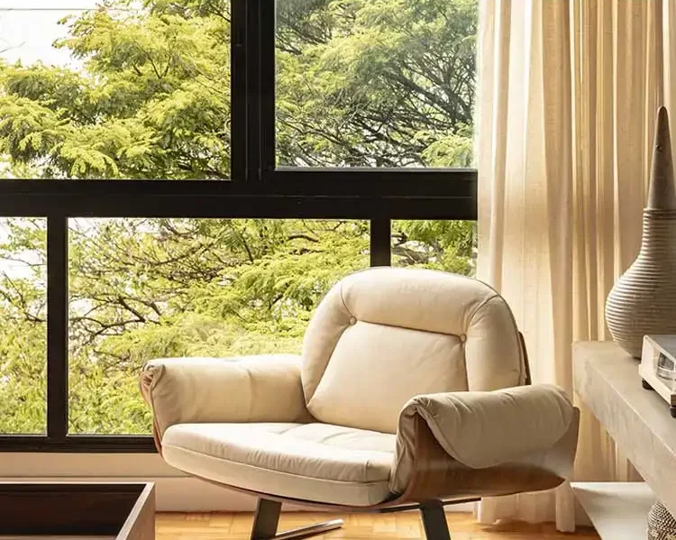 Beige armchair in a room with large windows showing greenery outside.